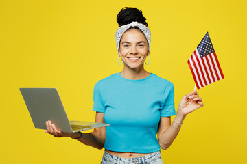 Wall Mural - Young happy Latin IT woman she wears blue top t-shirt headband casual clothes hold American flag use work on laptop pc computer isolated on plain yellow background studio portrait. Lifestyle concept.