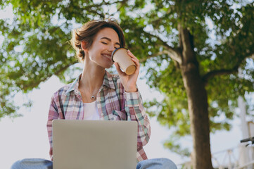 Wall Mural - Close up young smiling woman wear shirt casual clothes work hold use laptop pc computer drink coffee sits walk rest relax in spring sunshine green city park outdoors on nature. Urban leisure concept.