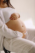 © Olena Zelena - Close up photo of pregnant woman in white clothes caressing her belly, seated on a chair indoors with copy space for tex