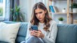© Four888 - Young woman sitting on couch, looking distressed and holding mobile phone, staring at screen with concern, her brows furrowed and lips pinched in worry.