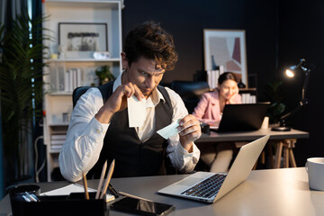 Sitting man with serious face tearing failure paperwork of business project at night lighting time at modern office with blurry secretary checking paper report with folder background. Postulate.