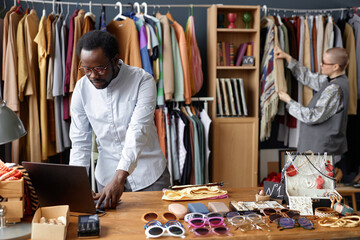 Wall Mural - Medium shot of African American salesman using laptop on wooden table with fashion accessories for sale at thrift store checkout, copy space