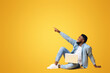 © Prostock-studio - Look there. African american guy sitting on floor with laptop and pointing aside at copy space on yellow background