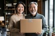 © kinara art design - A happy elderly couple holding a cardboard box, smiling in a cozy indoor setting.