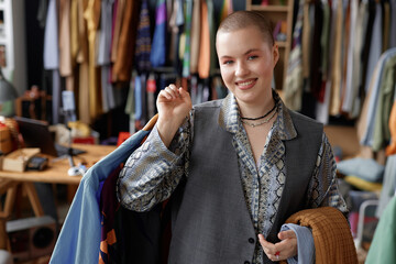 Waist up portrait shot of young smiling fashionable woman with buzz cut dressed in vintage clothes holding second hand sweaters while looking at camera at thrift store, copy space