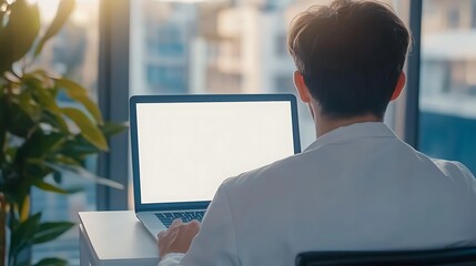 Wall Mural - Man using laptop with a blank screen, sitting in front of a window with a view of the city.