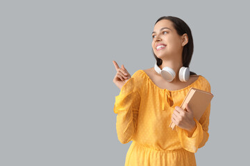 Canvas Print - Happy young woman with headphones and book pointing at something on grey background