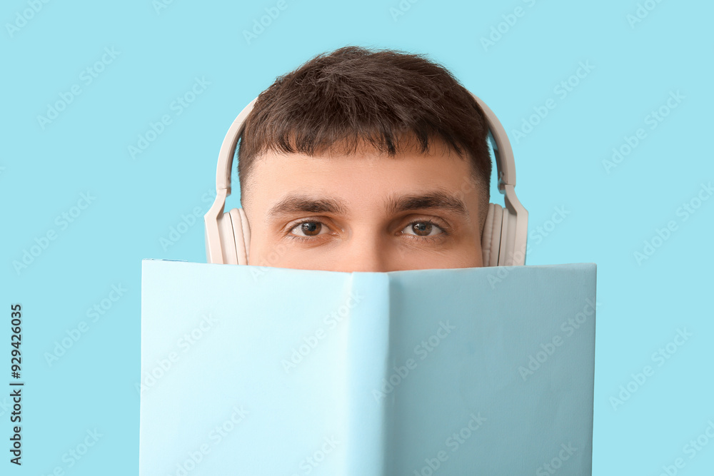 Handsome young man in headphones with book on blue background, closeup. Concept of audiobook
