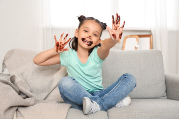 Sticker - Funny little Asian girl with chocolate on her hands sitting on sofa at home