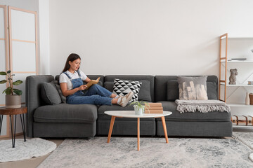 Poster - Beautiful young woman reading book and sitting on black sofa at home