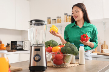 Poster - Young Asian woman with fresh vegetables and blender at table in kitchen