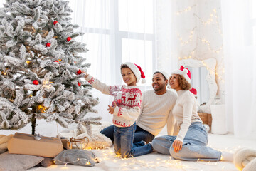 African American family in Santa hat celebrate New Year together at home and decorate Christmas tree, teenage boy celebrates New Year with mom and dad and holds red toy