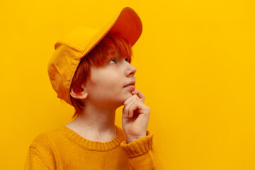 thoughtful red-haired teenage boy in cap planning and thinking over yellow isolated background, serious child in hat with orange hairstyle dreaming and imagining