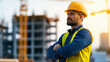 © khonkangrua - A construction worker wearing a hard hat and safety vest stands confidently at a building site with structures in the background.