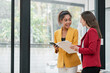 © Satori Studio - Two businesswomen in colorful blazers discussing work in a bright, modern office with large windows and greenery outside.
