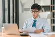 © phaitoon - A man wearing a blue shirt and a tie is sitting at a desk with a laptop open in front of him. He is looking at a piece of paper and he is focused on the task at hand