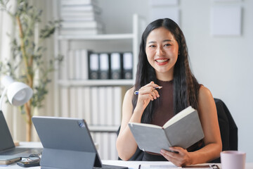 Asian businesswoman is smiling while holding a pen and an open notebook in a modern office