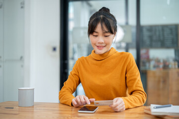 Wall Mural - A beautiful Asian woman holding a credit card and using her smartphone, paying bills online.