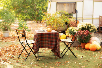 Cozy autumn atmosphere in garden - table with tablecloth and chairs, flowers, fallen leaves, pumpkins. Trailer with fall decor. Backyard with orange pumpkin. RV house porch with garden furniture.	
