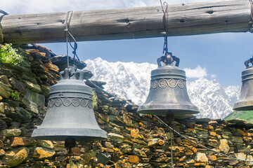 Two bells hang on a wooden log near a stone wall in the courtyard of the Lamaria Monastery in the village of Ushguli. The snow capped peaks of Mount Shkhara are in the background.