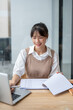 © bongkarn - A beautiful Asian businesswoman working on her laptop and business paperwork at a table in a room.