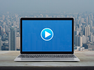 Canvas Print - Play button on modern laptop computer screen on wooden table over city tower and skyscraper, Business music online concept