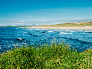 Wall Mural - Green grass grows on cliff and amazing waves and sandy Bundoran beach in Ireland on warm sunny day. Popular tourist and surfers area. Blue cloudy sky. Irish landscape. Rich saturated color.