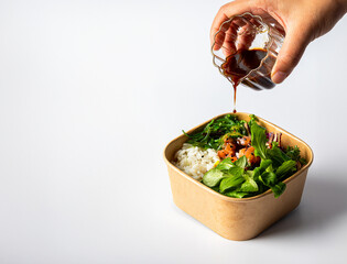 A woman's hand pours sauce over a healthy poke bowl with rice, algae, bites of salmon, and fresh vegetables