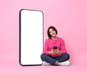 Poster - Beautiful young woman with smartphone sitting near big mobile phone with blank screen on pink background