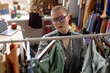 © Seventyfour - Portrait shot through clothing rack of fashionable female customer in vintage outfit and trendy aviator glasses choosing second hand clothes at thrift store, copy space