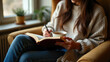 © SerPak - A woman in a cozy beige sweater sits comfortably in a chair, reading a book and holding a cup of tea, beside a window with potted plants and natural light pouring in.