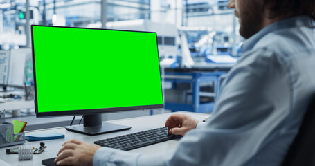 Fototapete - Young Industrial Software Engineer Working on a Desktop Computer with Green Screen Mock-Up Display. Modern Factory with High-Tech Machinery and Robotic Arms in the Assembly Hall