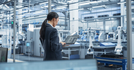 Fototapete - Southeast Asian Engineer Using Laptop Computer and Looking Around a Factory Facility with Equipment Producing Modern Electronic Components for Different Industries. Footage From the Back
