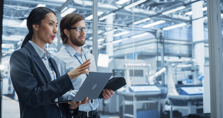 Fototapete - Two Multiethnic Engineers Standing in Office, Using Laptop and Tablet Computers at an Electronics Factory. Machines are Undergoing Maintenance, and Specialists Monitoring the Progress on Devices