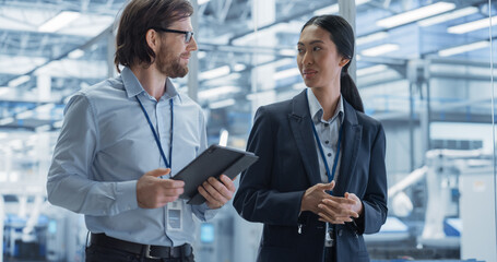 Leinwandbilder - Two Diverse Engineers Walking in Office, Using Tablet Computer at a Modern Electronics Factory. Female and Male Specialists Talking, Monitoring Robots on Modern Assembly Line.