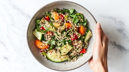 A bowl of quinoa salad with organic vegetables, featuring a hand holding the bowl in a simple, clean composition with a white background.