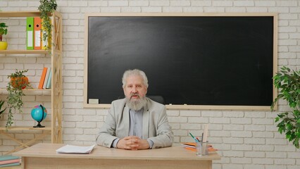 Aged man college professor sit at desk in classroom in front of chalkboard talking to students, explains lesson. Education concept.