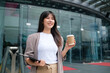 © wattana - Businesswoman standing outside a modern office building, holding a clipboard in one hand and a takeaway coffee cup in the other.