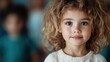 © Dulemegapixel - A young girl with curly hair and a thoughtful expression looks directly at the camera, with other children slightly out of focus in the background. Innocence and contemplation.