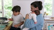 © Marco - Mother holding baby while sitting next to an older child eating at the dining table. The scene reflects the multitasking and nurturing aspects of parenthood in a cozy home environment