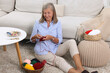 © New Africa - Smiling senior woman knitting near sofa at home
