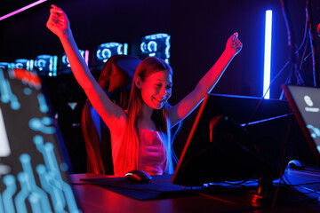 Happy young girl is playing online video games on computer, she raises her hands up and rejoices.