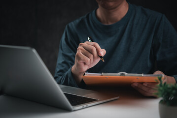 Wall Mural - Businessperson writing on a clipboard while working on a laptop, representing planning and organization. Suitable for project management content