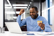 © Liubomir - African American mature businessman rejoicing successful online transaction at office. Smiling man using laptop and holding credit card, experiencing moment of achievement.