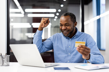 African American mature businessman rejoicing successful online transaction at office. Smiling man using laptop and holding credit card, experiencing moment of achievement.