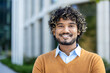 © Liubomir - Confident young professional with curly hair and warm smile standing outdoors near modern office building. Image conveys success, positivity, and approachable demeanor in business context