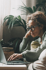 Close-up of a young woman lying on a cozy sofa at home, browsing the internet on her laptop with a slightly bored expression, searching for content to pass time, in a relaxed and casual atmosphere