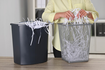 Wall Mural - Woman putting shredded paper strips into trash bin at wooden table in office, closeup