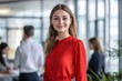 © Elmira - A young woman dressed in a vibrant red blouse smiles confidently in a contemporary office filled with colleagues engaged in work-related activities