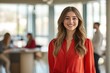 © Elmira - A confident young woman in a red blouse is smiling warmly in a bright office with colleagues busy in the background, creating a lively work atmosphere